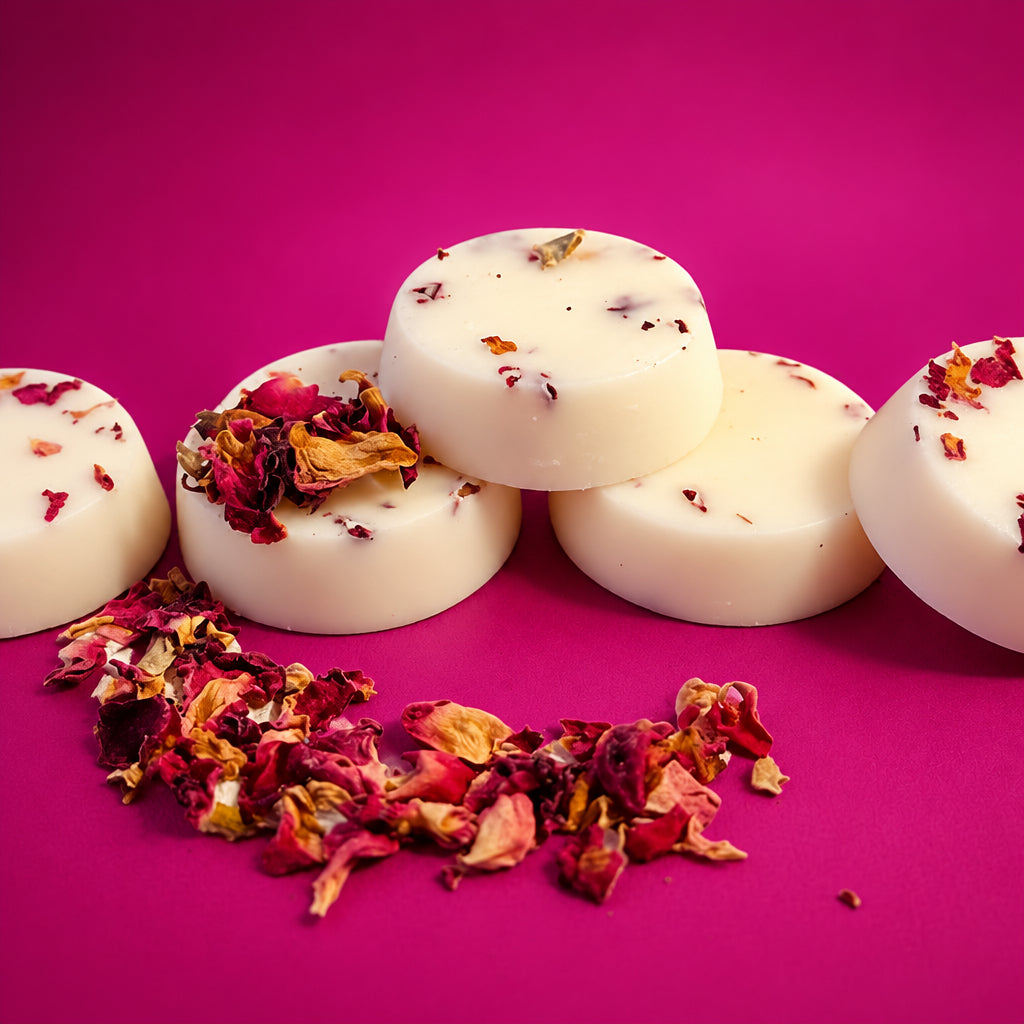 Three round off-white soap bars with dried rose petals embedded, arranged on a white surface with scattered dried red and pink rose petals in the foreground.