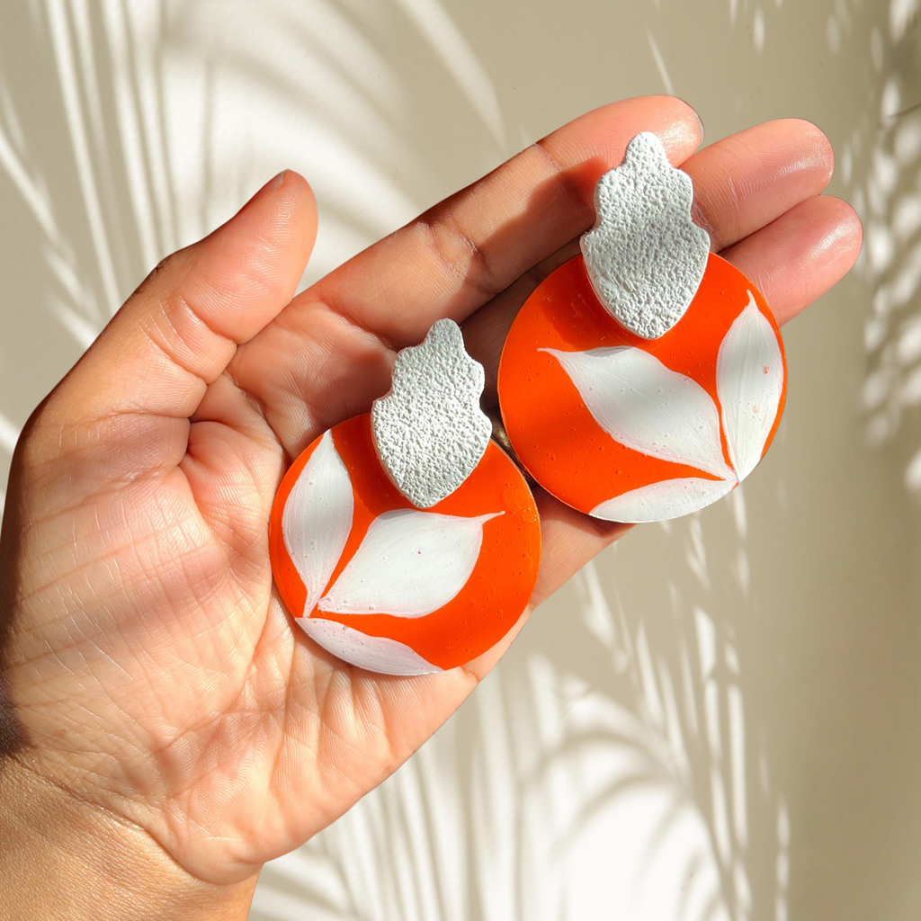 Hand holding two orange and white leaf-shaped earrings against a neutral background