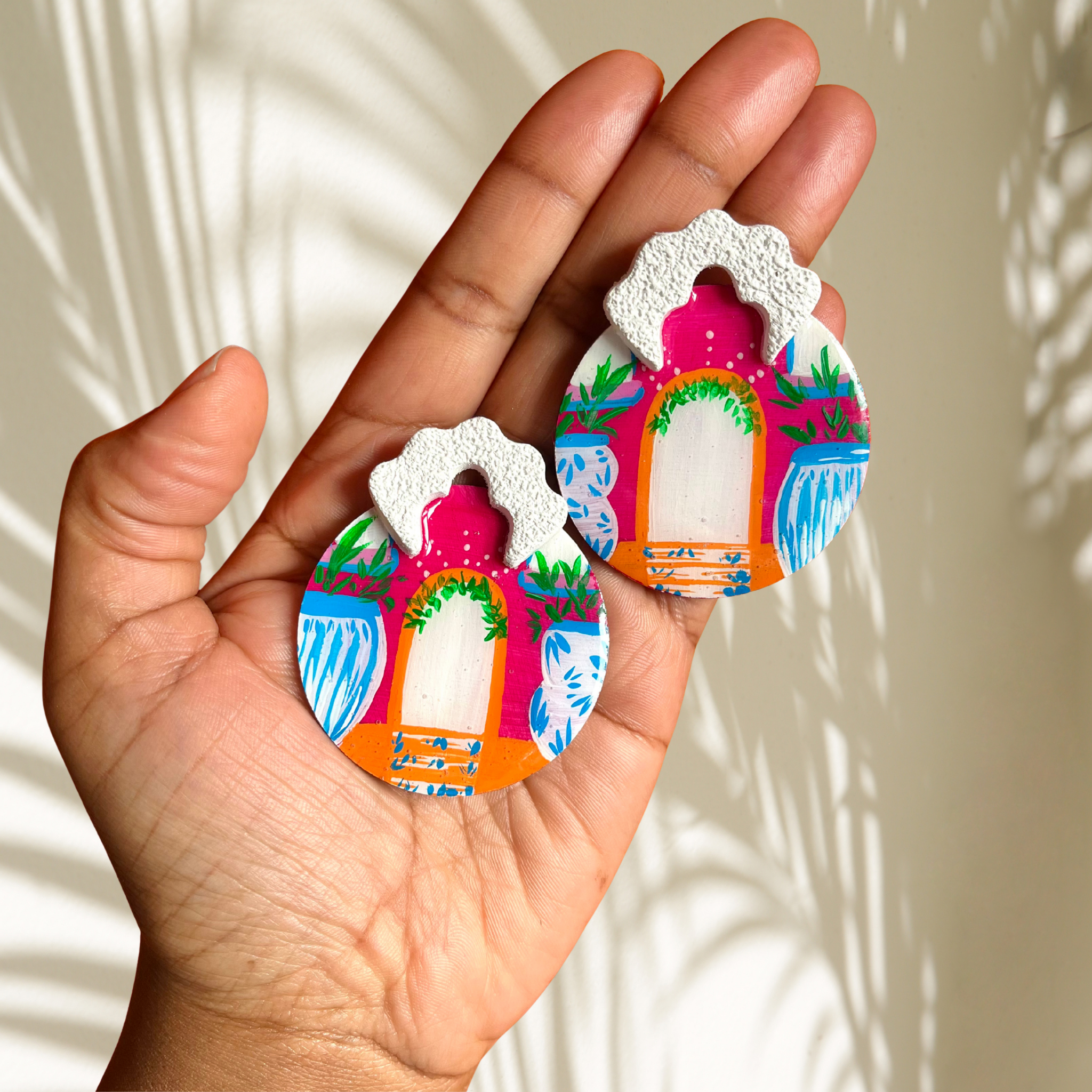 Hand holding two colourful earrings with a decorative design of a little house against a neutral background