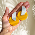 Hand holding two yellow and stone imitation earrings against a neutral background
