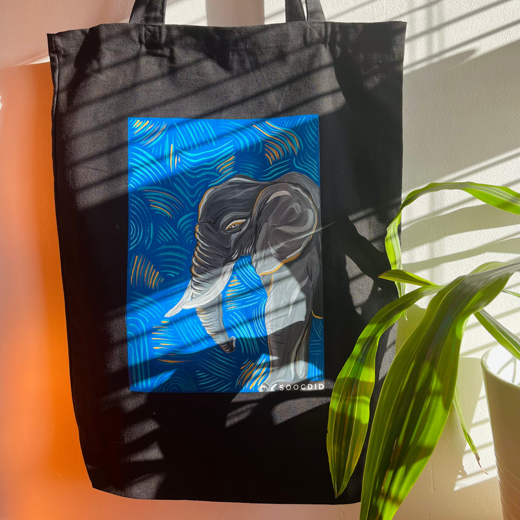 Black tote bag with a colourful elephant design on a plant and light-coloured blue wall background