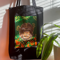 Black tote bag with a colorful monkey illustration on a green background, held by a person with a plant in the foreground.
