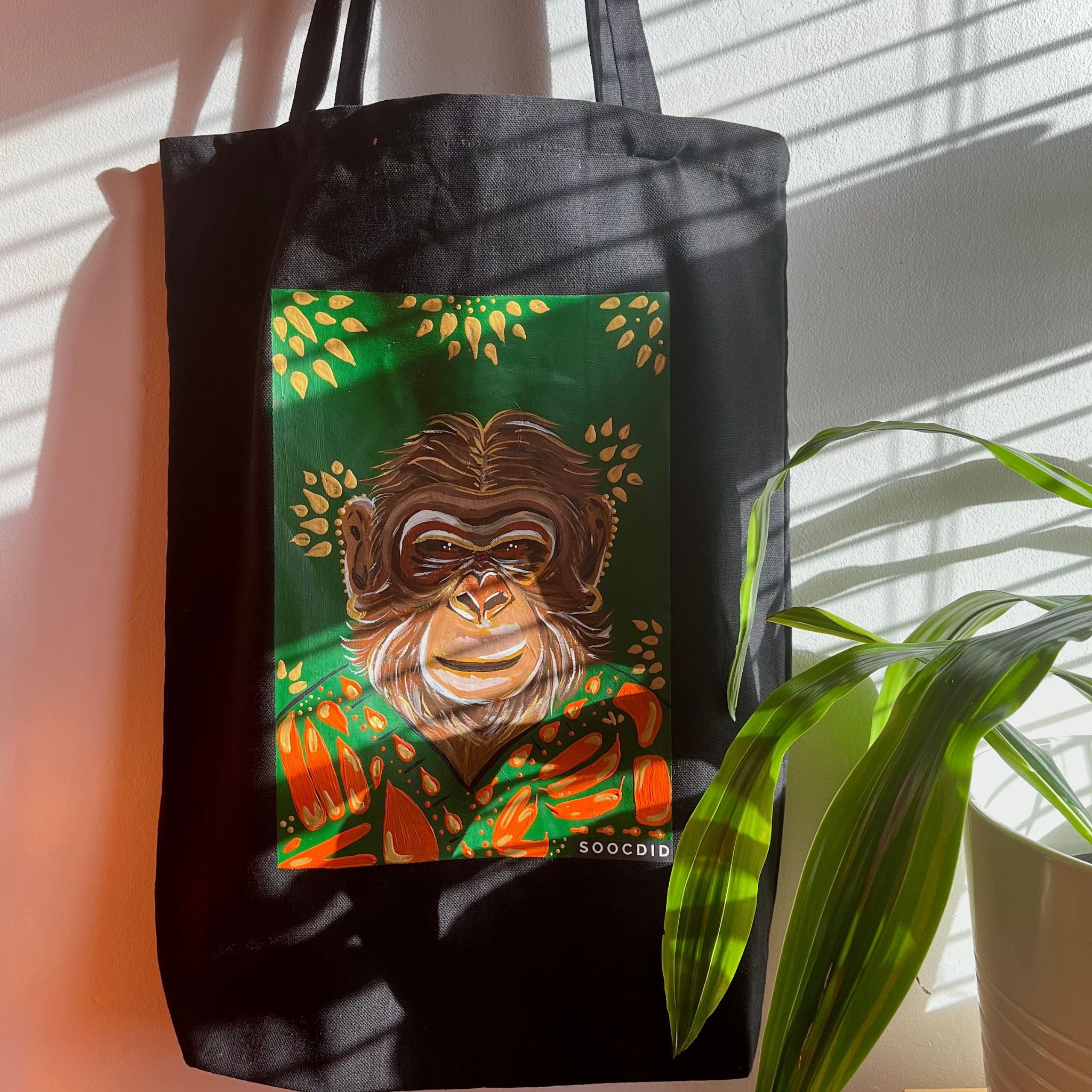 Black tote bag with a colorful monkey illustration on a green background, held by a person with a plant in the foreground.